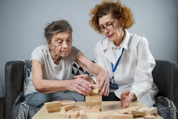 Jenga game. Theme is dementia, aging and games for old people. Caucasian senior woman builds tower of wooden blocks with the help of a doctor as part of a therapy and jenga game at a patient\'s home.