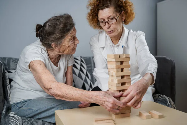 Jenga game. Theme is dementia, aging and games for old people. Caucasian senior woman builds tower of wooden blocks with the help of a doctor as part of a therapy and jenga game at a patient's home.