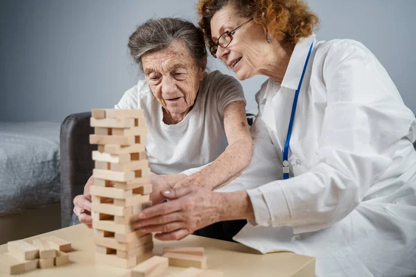 Jenga oyunu. Temaları bunama, yaşlanma ve yaşlılar için oyunlar. Kafkasyalı son sınıf öğrencisi kadın, bir hastanın evindeki terapi ve jenga oyununun bir parçası olarak bir doktorun yardımıyla ahşap bloklardan kule inşa ediyor..