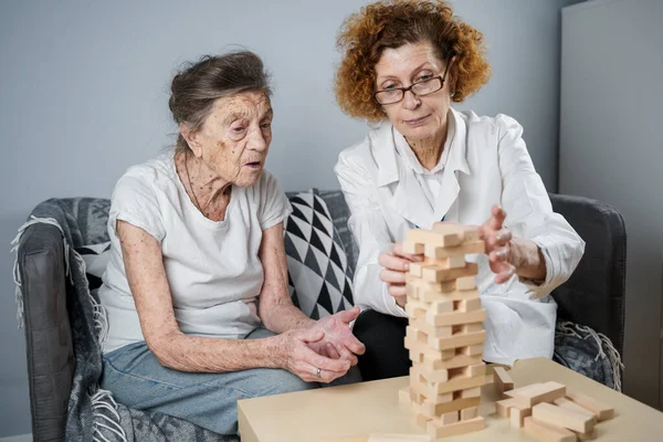 Jenga game. Theme is dementia, aging and games for old people. Caucasian senior woman builds tower of wooden blocks with the help of a doctor as part of a therapy and jenga game at a patient\'s home.