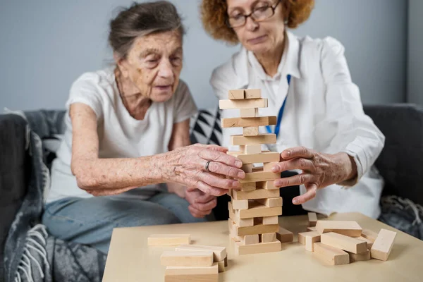Jenga oyunu. Temaları bunama, yaşlanma ve yaşlılar için oyunlar. Kafkasyalı son sınıf öğrencisi kadın, bir hastanın evindeki terapi ve jenga oyununun bir parçası olarak bir doktorun yardımıyla ahşap bloklardan kule inşa ediyor..