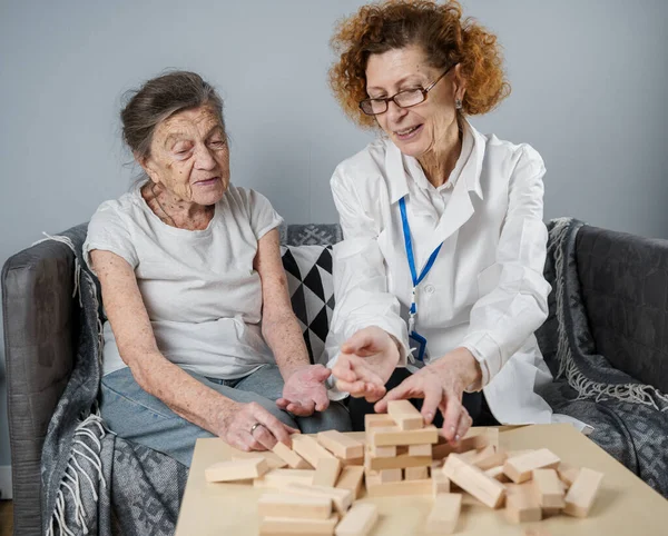 Jenga oyunu. Temaları bunama, yaşlanma ve yaşlılar için oyunlar. Kafkasyalı son sınıf öğrencisi kadın, bir hastanın evindeki terapi ve jenga oyununun bir parçası olarak bir doktorun yardımıyla ahşap bloklardan kule inşa ediyor..