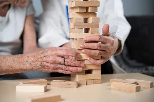 Mature doctor conducting session, therapy for senior patient in nursing home, training fine motor skills for dementia, alzheimer disease and recovery institute by folding wooden blocks, playing jenga.