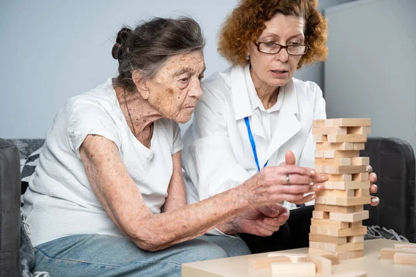 Jenga game. Theme is dementia, aging and games for old people. Caucasian senior woman builds tower of wooden blocks with the help of a doctor as part of a therapy and jenga game at a patient\'s home.