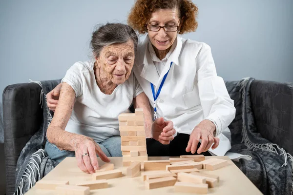 Senior woman practice skills, build wooden blocks, build tower and try not to let it fall, Jenga game. Old patient pull out block, place on top, support doctor during therapy dementia in house.