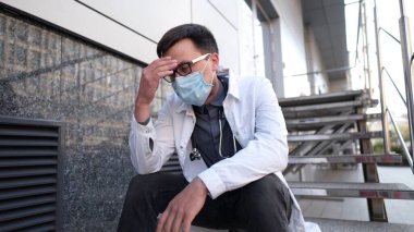 Kafkasyalı genç doktor, klinik binasının yanındaki merdivenlerde oturur, yorgun ve mutsuzdur, burnunu ve gözlerini ovuşturur, yorgun ve baş ağrısı hisseder. Sağlık çalışanı stresi ve hüsran konsepti.