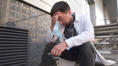 Hastanede uzun bir vardiyadan sonra merdivenlerde dinlenen ciddi bir doktor. Maskeli yorgun doktor, beyaz üniformalı ve steteskoplu. Kliniğin dışındaki merdivenlerde dinleniyor. Stres baş ağrısı ve gözlerini ovuşturuyor..
