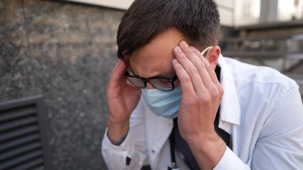 Worried and tired doctor sitting on stairs outside hospital. Exhausted physician in white lab coat and medical protective mask, sit on staircase clinic. Stress healthcare worker during COVID-19.