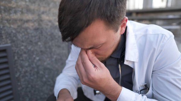 Caucasian young doctor man sits down on the stairs near the clinic building, tired and unhappy rubbing his nose and eyes, feeling tired and headache. Health care worker stress and frustration concept.
