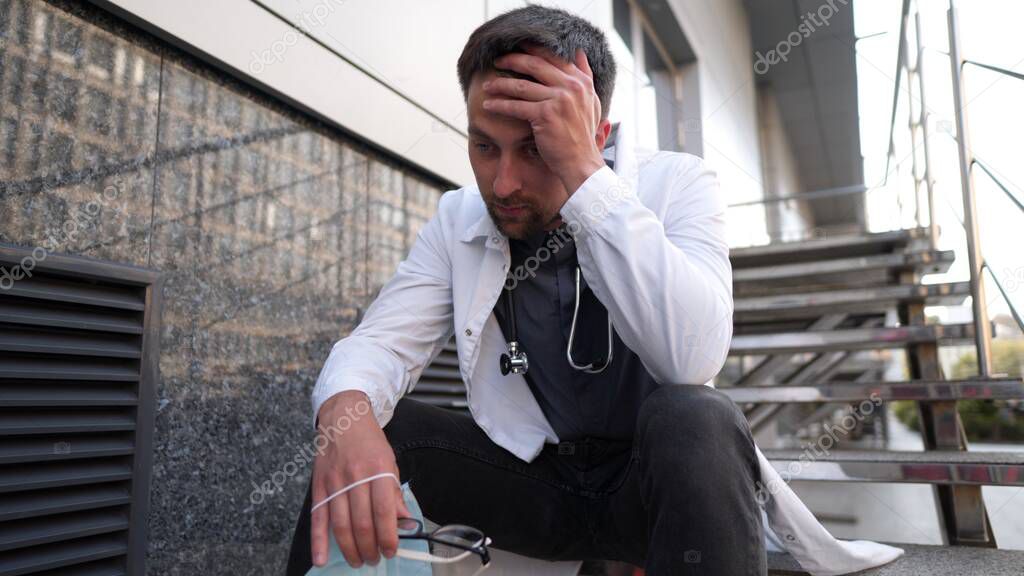 Worried and tired doctor sitting on stairs outside hospital. Exhausted physician in white lab coat and medical protective mask, sit on staircase clinic. Stress healthcare worker during COVID-19.