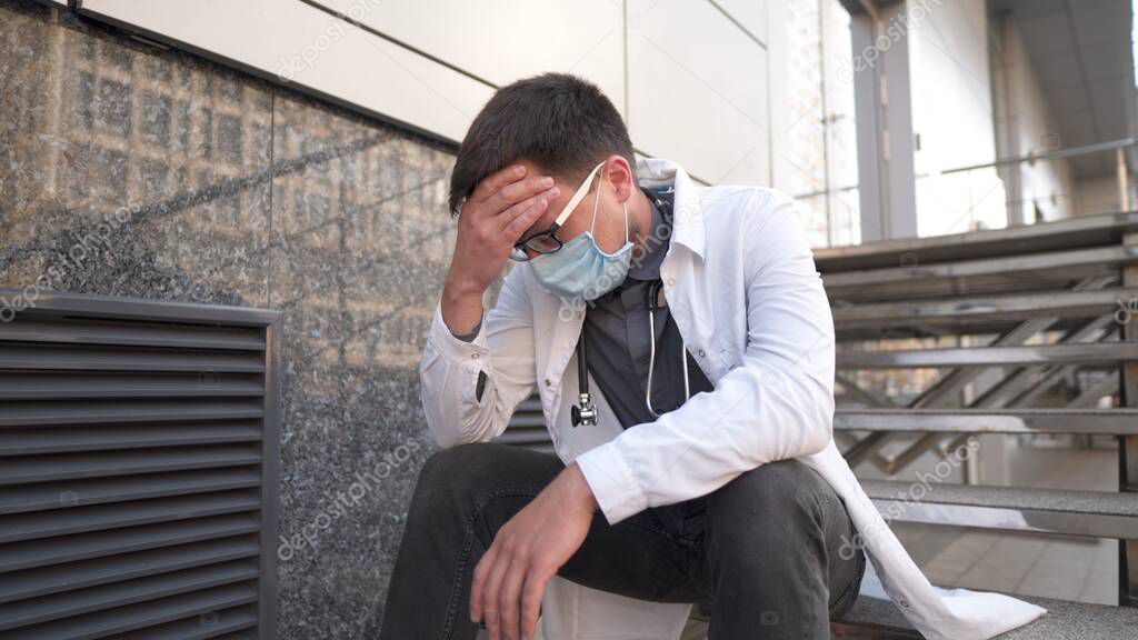 Caucasian young doctor man sits down on the stairs near the clinic building, tired and unhappy rubbing his nose and eyes, feeling tired and headache. Health care worker stress and frustration concept.