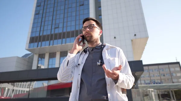 Young Caucasian doctor in white medical coat consults patient on cell phone during break outside hospital. Medical worker talking on mobile phone near clinic. Physician in uniform talk on phone.