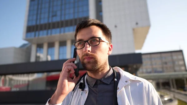 Portrait of tired exhausted doctor having phone call outside hospital in morning. COVID-19, Coronavirus pandemic. Medical practitioner talk on cellphone near clinic. Calling Out. Dr talking on phone.