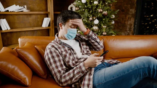 Sad man in mask surfing Internet alone at home on couch at Christmas time. Quarantine during New Year holidays ruined mood and introduced into depression, communication with friends and family online.