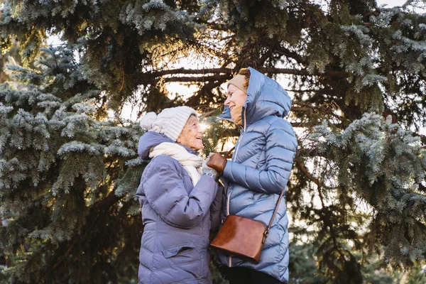 Concept of happy family, old age, emotions, senior care in retirement age. Active senior grandmother and adult daughter hugging outside in winter against background of Christmas tree in forest.