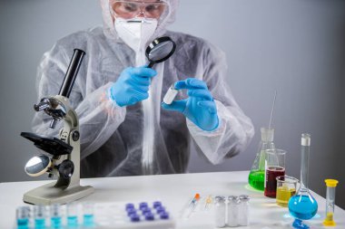 Tehlikeli madde giysisi ve gözlüklü bilim adamları aşı şişesi ve büyüteç tutuyor, bakıyor ve araştırıyor. Epidemiyoloji virüs testi. İlaç araştırmaları, klinik deneyler. Biyoteknoloji kimyageri laboratuvarda..