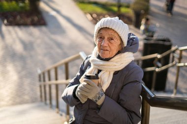 Elinde kahve tutan neşeli yaşlı bayan, şehirde kışlık kıyafetler giyiyor. Kahveli güzel yaşlı bayan. Yaşlılıkta aktif dinlenme, yürüyüş sırasında kahve molası. Sıcak içecek, 90 yıl emekli..