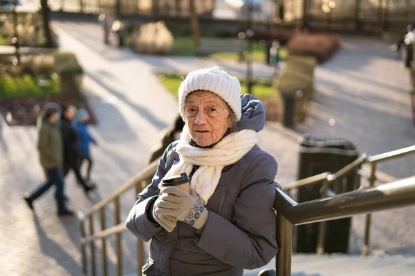 冬の服 白いニット帽 晴れた天気の街でスカーフに行くためにコーヒーを飲みながら幸せなシニア白人女性の肖像画 コーヒーを屋外で高齢者の女性 楽しみ — ストック写真