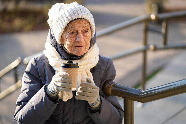 Joyful senior lady holding coffee to go, dressed winter clothes in city. Beautiful elderly lady with cup coffee to go. Active rest in old age, coffee break during walk. Hot drink, retired 90 years.