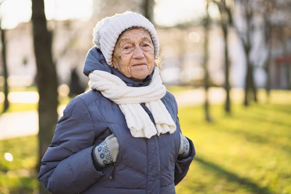 Portrait Caucasian senior woman with gray hair and deep wrinkles 90 years old posing in warm clothes, white knitted scarf and hat in park, sunny frosty weather. Active old age, walking elderly female.