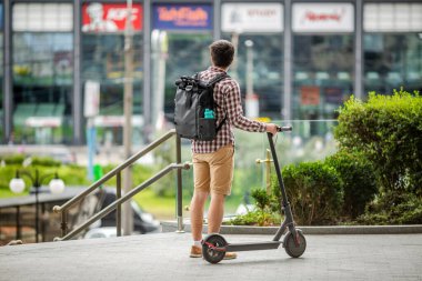 Elektrikli scooter 'ı olan genç beyaz bir adam, kürsünün üzerinde duruyor, büyük modern şehirde trafik sıkışıklığını atlatarak en uygun rotayı planlıyor. Tema araba ve şehir taşımacılığına alternatif, kişisel hareketlilik.
