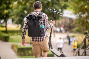 Kurye elektrikli scooter 'ı merdivenlerden araba parkına taşıyor. Sırtında siyah bir sırt çantası olan sıradan giyinmiş genç adam şehri hızlıca dolaşmak için alternatif kişisel e-ulaşım araçları kullanıyor..