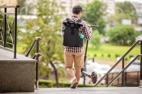 Courier carries an electric kick scooter down the stairs to car parking. Casual dressed young man with a black backpack on back uses alternative personal e-transport to get around the city quickly.