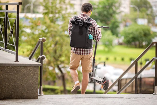 Adult man lifts electric scooter and holds it in hands while descending stairs at public space. Theme inclusiveness and barrier-free urban space, problems of transport infrastructure and movement.