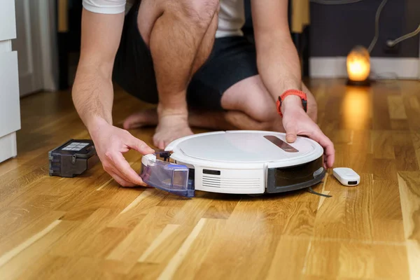 Caucasian young man doing white robot vacuum cleaner maintenance. Autonomous wireless modern vacuum cleaner. Male repairing robot cleaner home on floor. Vacuum cleaner robot repair concept.