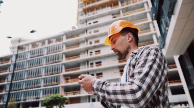 İnşaatın kabulü işe yarıyor. Kasketli bir adam telefonda bir inşaat planı tutuyor. Adam bina yapısının uygunluğunu kontrol ediyor. İnşaatın kalitesinin değerlendirilmesi.
