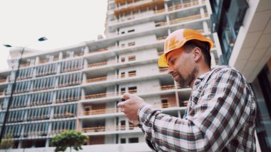 İnşaatın kabulü işe yarıyor. Kasketli bir adam telefonda bir inşaat planı tutuyor. Adam bina yapısının uygunluğunu kontrol ediyor. İnşaatın kalitesinin değerlendirilmesi.