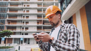 İnşaatın kabulü işe yarıyor. Kasketli bir adam telefonda bir inşaat planı tutuyor. Adam bina yapısının uygunluğunu kontrol ediyor. İnşaatın kalitesinin değerlendirilmesi.