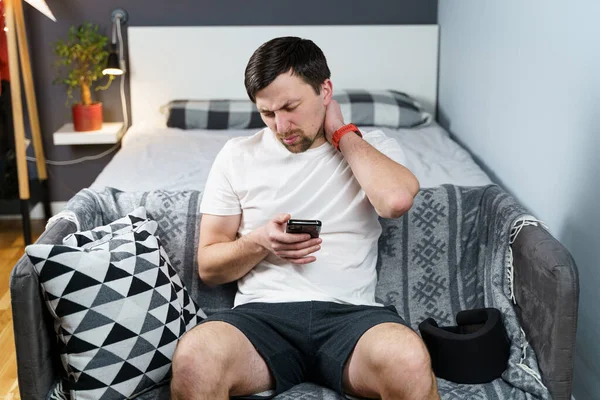 Young man after car accident suffering at home. Male in foam cervical collar reading message on cellphone, feeling pain in neck. Guy wearing neck brace to support neck at home on couch.