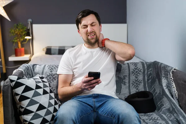 Caucasian male has neck injury to relieve pain, puts on cervical brace made of soft black cotton, male uses smartphone at home sitting on couch in cervical collar, cervical brace relieves tension.