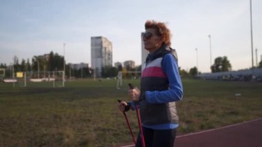 Spor sahasında yürüme direğiyle çalışan yaşlı bir kadın. Yaşlılar sağlıklı bir yaşam tarzı. Kıdemli İskandinav yürüyüşü. Şehir stadyumunda koşan bastonlu olgun bir kadın.