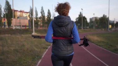 Spor sahasında yürüme direğiyle çalışan yaşlı bir kadın. Yaşlılar sağlıklı bir yaşam tarzı. Kıdemli İskandinav yürüyüşü. Şehir stadyumunda koşan bastonlu olgun bir kadın.