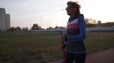 Spor sahasında yürüme direğiyle çalışan yaşlı bir kadın. Yaşlılar sağlıklı bir yaşam tarzı. Kıdemli İskandinav yürüyüşü. Şehir stadyumunda koşan bastonlu olgun bir kadın.