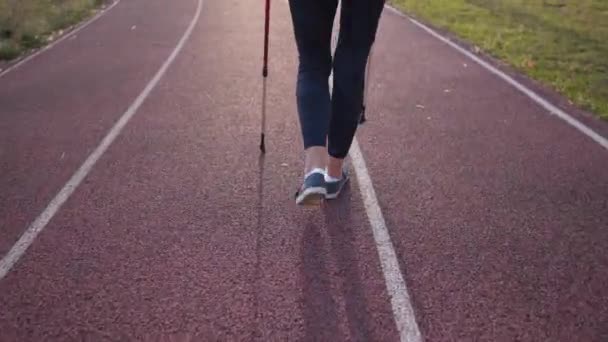 Concepto de estilo de vida saludable. Mujer caucásica madura haciendo ejercicio cardiovascular caminando con palos o caminando nórdico en pista de estadio en la ciudad. Actividad de los ancianos. Mujer mayor caminando con bastones — Vídeos de Stock