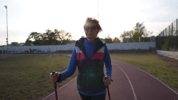 Senhora envelhecida trabalhando fora com postes de passeio no sportsground. Idosos estilo de vida saudável. Caminhada nórdica. Mulher madura com bengalas ao ar livre na pista de corrida do estádio na cidade — Vídeo de Stock