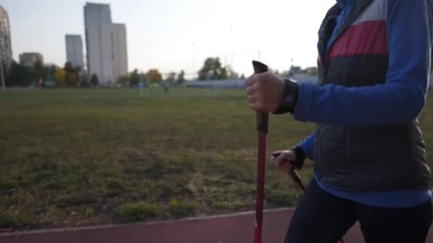 Madurez por edad, estilo de vida activo y bienestar. Mujer jubilada alegre con bastones alrededor del estadio de la ciudad. Mujer madura haciendo Nordic walking outdoor. Anciana deportista con bastones de trekking a pie — Vídeos de Stock