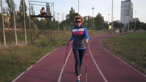 Mujer mayor entrenando con bastones en el estadio. Las hembras maduras hacen ejercicios de caminata nórdica en pista de atletismo. Concepto de estilo de vida saludable. Ejercicio femenino activo con bastones de trekking en la ciudad — Vídeos de Stock