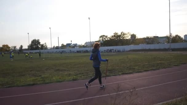 Dame âgée travaillant avec des bâtons de marche sur terrain de sport. Personnes âgées mode de vie sain. Marche nordique senior. Femme mûre avec des bâtons de marche à l'extérieur sur la piste de course du stade en ville — Video