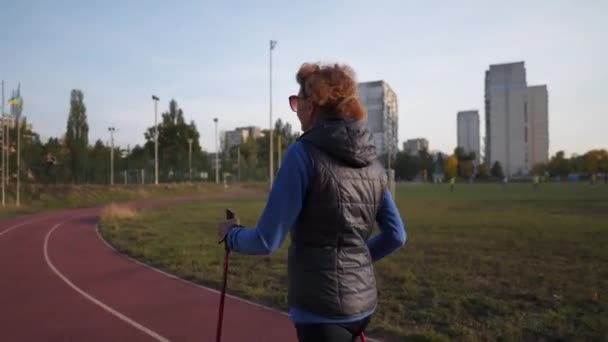 Starší žena trénuje s chodícími tyčemi na stadionu. Starší ženy dělají Nordic walking cvičení na běžecké dráze. Zdravý životní styl. Aktivní ženské cvičení s trekingovými holemi ve městě — Stock video