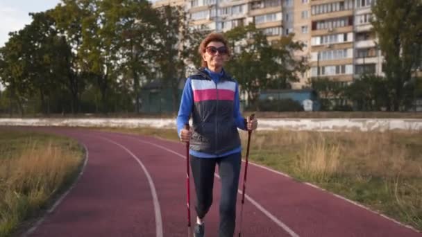 Aged lady working out with walking poles on sportsground. Elderly people healthy lifestyle. Senior nordic walking. Mature woman with walking sticks outdoors on running track of stadium in city — Stock Video