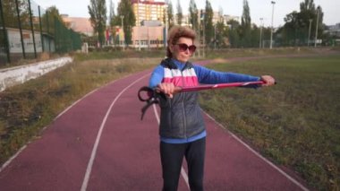 Kıdemli kadın, stadyumun koşu pistinde yürüyüş direğiyle yürümeden önce geriniyor. Sportif yaşlı kadın antrenman çubuklarını ısıtıyor. Aktif yaşam tarzı ve sağlık. Çeşitli kuzey kutupları alıştırmaları