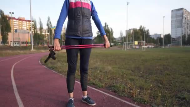 Atlétikai végzős nő testmozgást végez bottal a futópálya stadionban edzés előtt skandináv séta. Sportos életmód öregkorra. Idős nő edzés sétapóznákkal a szabadban — Stock videók