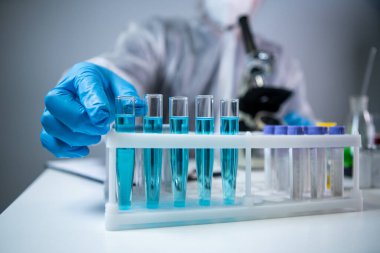 Koruyucu tulum giyen bilim adamı ya da doktor mavi reaktörlü test tüpü, kimyasal sıvı, mikroskop, laboratuvar biyokimyası içeren laboratuvar camları alıyor. Tıp ve araştırma konsepti