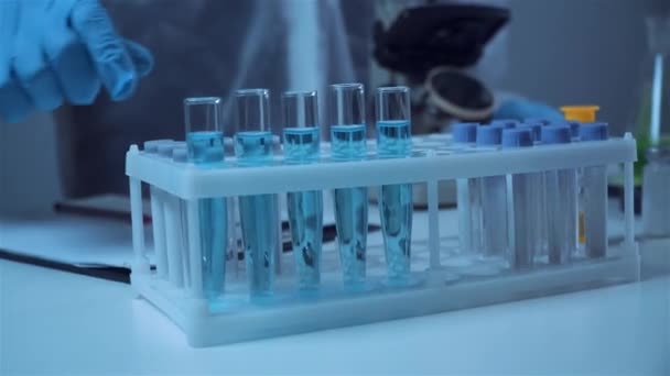 Recherche en laboratoire de biochimie, chimiste en combinaison de protection analysant un échantillon bleu en laboratoire avec équipement, microscope et verrerie d'expériences scientifiques contenant du liquide chimique — Video