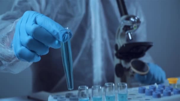 Wetenschapper of arts in beschermende overalls, neemt reageerbuis met blauw reagens, laboratorium glaswerk met chemische vloeistof, microscoop, laboratorium biochemie. Geneeskunde en onderzoek — Stockvideo