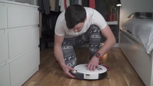 Ein junger Vater in Nachthemd mit Winterschmuck und weihnachtlichen Socken bedient einen Roboterstaubsauger, um das Haus für Neujahr und Weihnachten zu säubern. Neue Technologien für Haushalte — Stockvideo